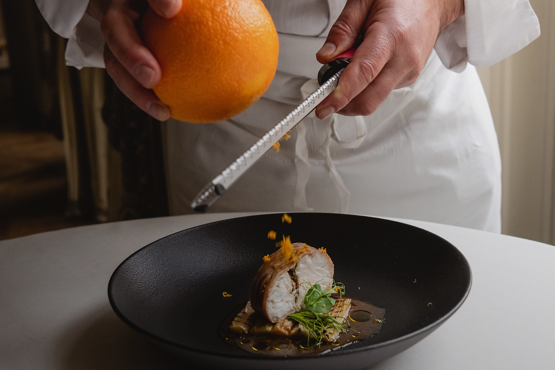Amelie-Poirier-Ritz Paris Design-1 Dans une ambiance lumineuse, une personne en veste de cuisine blanche râpe le zeste d'une orange au-dessus d'une assiette dressée dans une grande assiette noire. Le plat présente un poisson roulé, des légumes verts, des herbes et une sauce brune, tandis que les zestes d’orange tombent délicatement sur la préparation gastronomique. L’image met en avant le geste du chef, la précision et la délicatesse de la cuisine française raffinée.