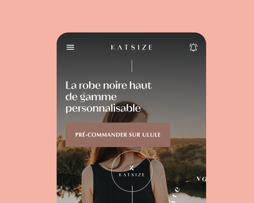Ecran mobile du site Katzsize affichant une femme en robe noire sur fond de paysage naturel, avec le texte 'La robe noire haut de gamme personnalisable'. Un bouton rose indique 'Pré-commander sur Ulule'. Le menu hamburger et une cloche de notification sont visibles en haut de l’interface.